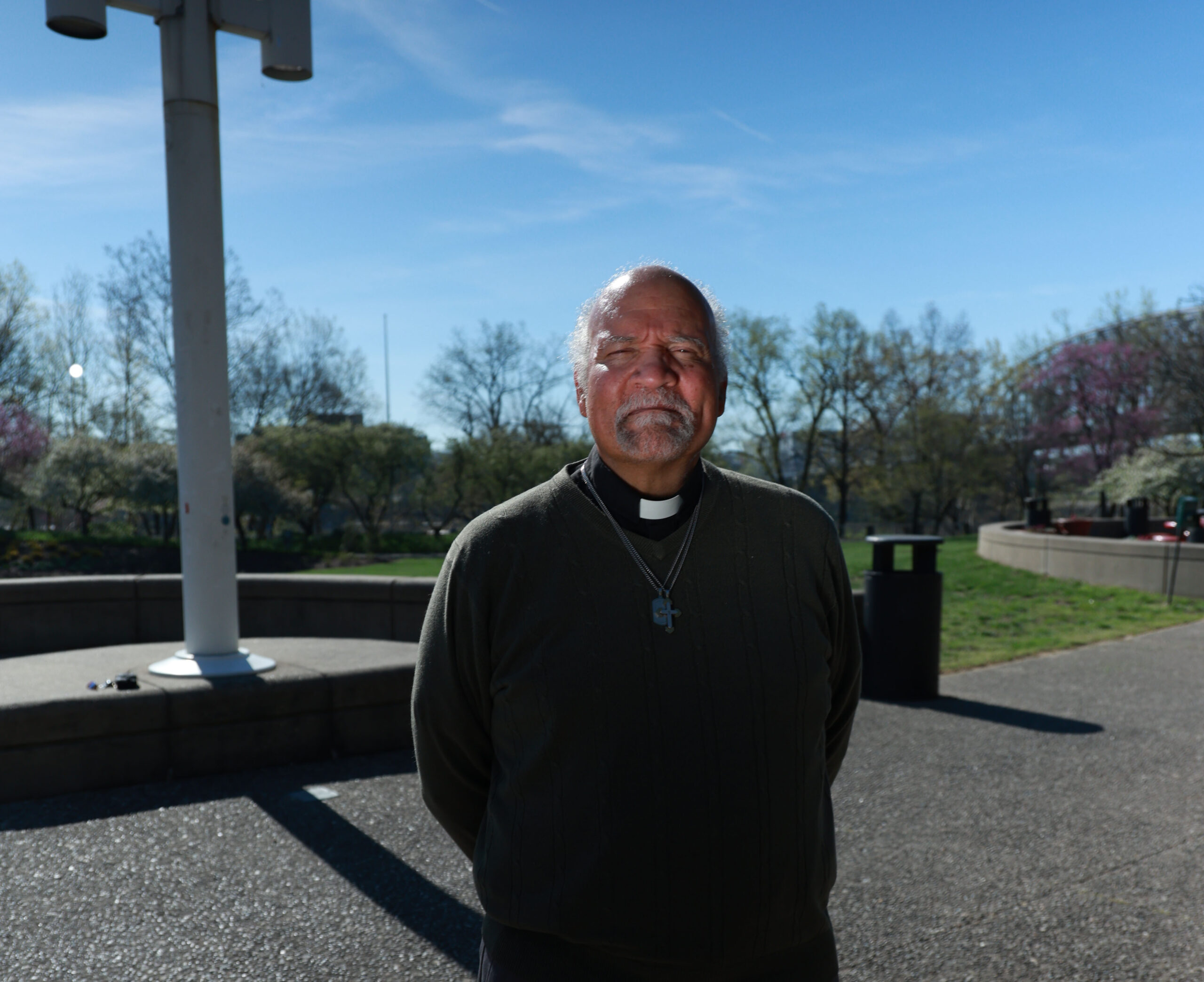 A man with a cross around his neck, standing in the sunlight outside.