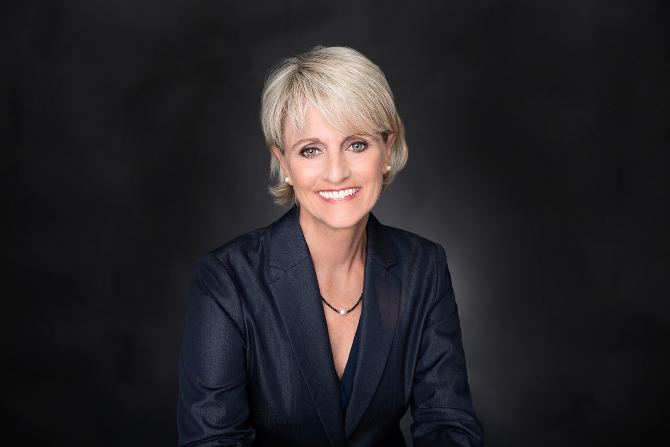 A white woman against a black background, with blond hair in a blazer smiling and looking into the camera