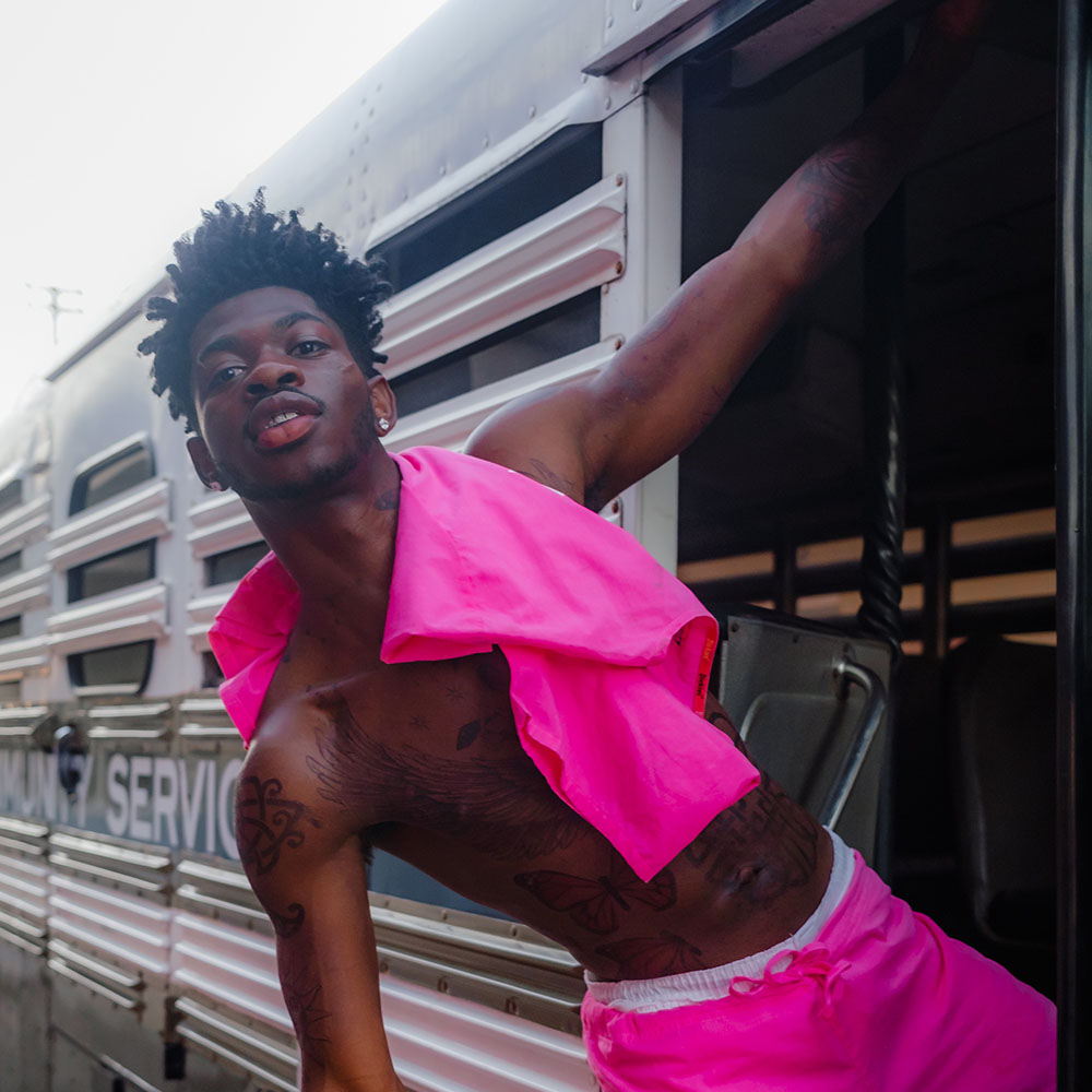 Lil Nas X, shirtless with a bright pink shirt over his shoulder and wearing bright pink shorts, leaning on the side of a train looking into the camera; credit Adrian Per